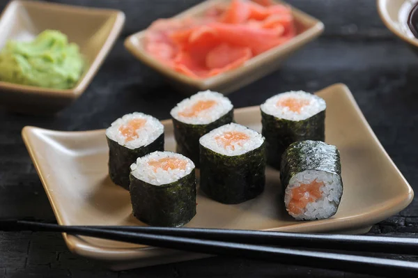 Roll with salmon on a light ceramic plate. Next to a plate of chopsticks. Ginger pickled and wasabi in bowls. Black wooden background. Close-up. Macro shooting.