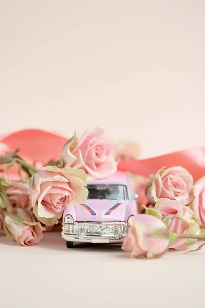 Pink toy car surrounded by a bouquet of spray roses. Free space to place text. The concept of holiday greetings. Light background. Vertical frame orientation.