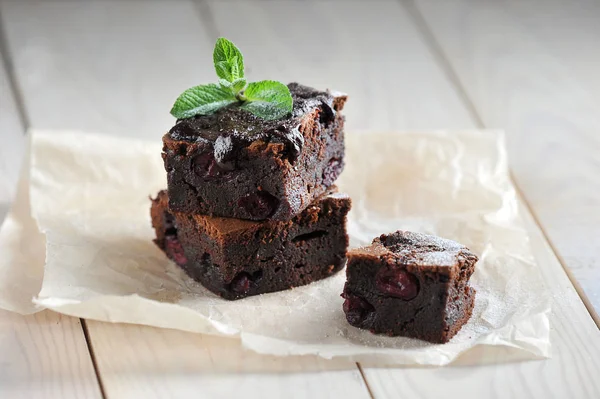 Chocolate brownie with cherry. Pieces of brownie laid out on paper. Light  wooden background. Close-up.