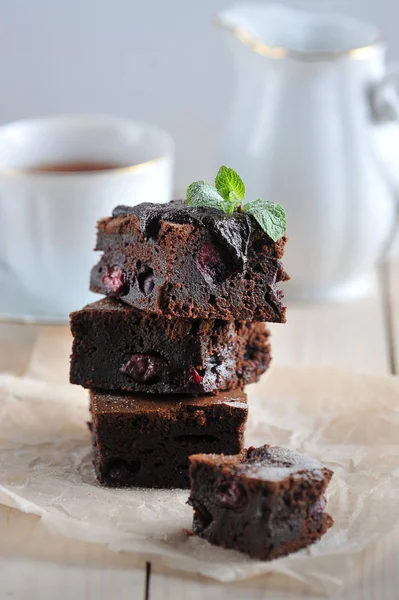 Chocolate brownie with cherry. Pieces of brownie laid out on paper. In the background is a cup with tea and a milkman. Light wooden background. Close-up. Vertical frame orientation.