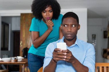 Jealousy african american woman checking chat on cellphone of boyfriend at home clipart