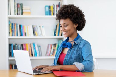 Beautiful african american young adult woman at computer indoors at desk at home clipart