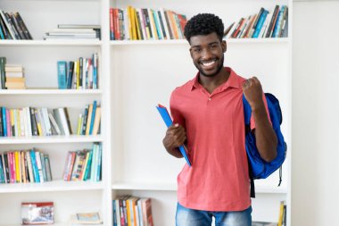Cheering afro american male student with backpack and copy space at library of university clipart
