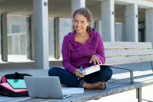 Pretty german female student learning with book and computer in front of school building outdoor in summer