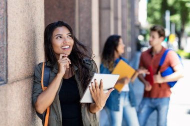 Tablet bilgisayarı ve arka planda arkadaşları olan genç Meksikalı öğrenci yaz aylarında şehir dışındaki üniversite binasının önünde.