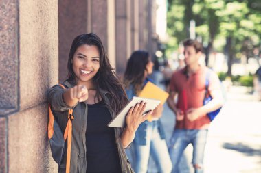 Güzel Latin Amerikalı bayan öğrenci dijital tablet bilgisayarıyla ve diğer genç yetişkinlerle üniversite binasının önünde.