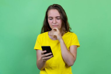 Esmer saçlı, genç bir Alman genç kadın yeşil arka planda izole edilmiş cep telefonuyla mesaj gönderiyor.