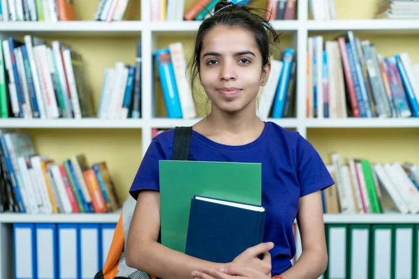 Üniversite kütüphanesinde kitap ve evrak işleriyle gülümseyen Hintli kız öğrenci.