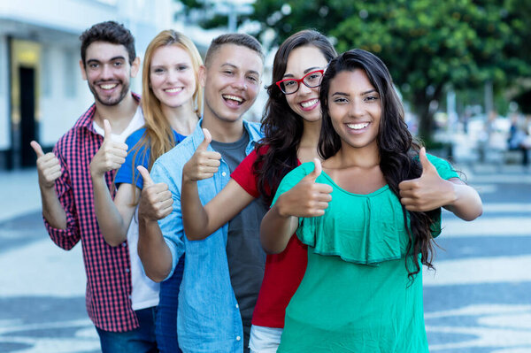 Group of five beautiful international young adults in line showing thumbs up outdoor in city