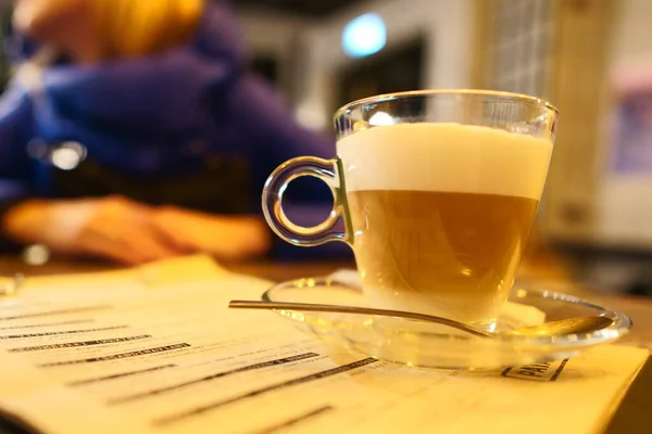 Taza de vidrio con café con leche en café sobre una mesa de madera vintage. Foto en luz tenue en colores cálidos en una mesa de madera con un menú bajo una taza de café, foto de estilo de vida —  Fotos de Stock