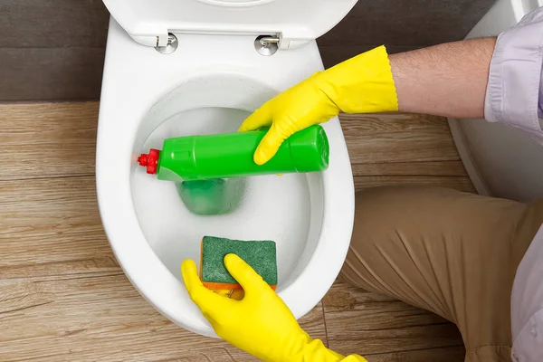 Ο άνθρωπος απολυμαίνει το κάθισμα της τουαλέτας ψεκάζοντας ένα πράσινο απολυμαντικό από ένα μπουκάλι. Ένας άντρας απολυμαίνει μια τουαλέτα στο σπίτι.. — Φωτογραφία Αρχείου