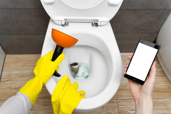 Overflowing broken toilet. clogged toilet. a smartphone with a white screen for advertising about plumbing. girl looking for help for an hour on the phone. mockup. — Stock Photo, Image