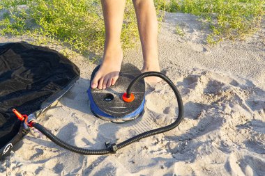A woman with air foot pump pumps an inflatable mattress or air bed at sandy beach. Foot inflates air mattress with foot pump on sand. clipart