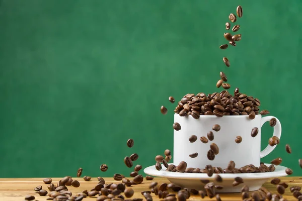 International day of coffee concept. close-up white coffee cup full of coffee beans. coffee beans fall into the white mug assign dark green background.