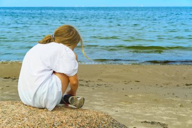 Sahildeki taşların üzerinde oturan ve denize bakan sevimli sarışın bir kızın arka tarafı. Hayal ya da Rahatlama konsepti.