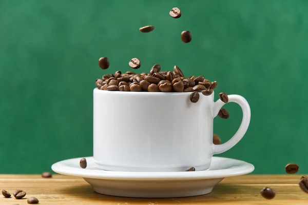 International day of coffee concept. close-up white coffee cup full of coffee beans. coffee beans fall into the white mug assign dark green background.