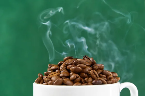 International day of coffee concept. close-up white coffee cup full of coffee beans on green background with smoke on top.