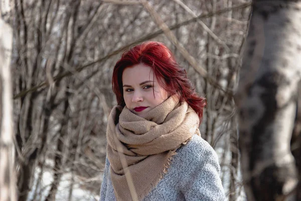 Pretty young women with red hair in beautiful beige scarf and grey coat posing in a winter forest or park. — Stock Photo, Image
