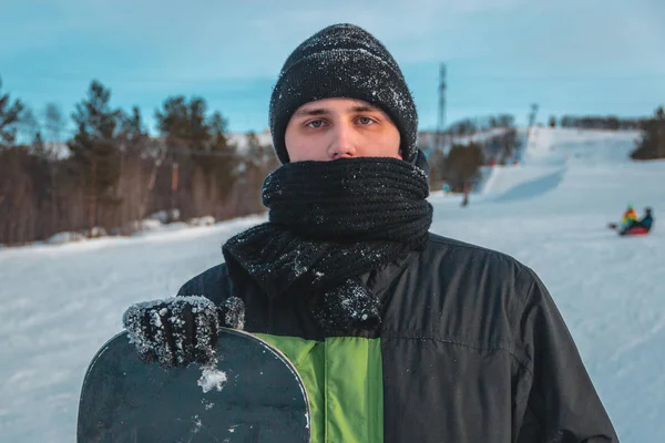 Snowboarder man with scarf in black hat make photoshoot on ski slope. On the background of ski slope. Close up view.