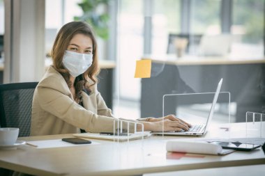 İş kadını yüz maskesi takıyor ve Coronavirus covid-19 salgınını korumak için masasında bölme kullanıyor. Sosyal ve iş mesafesi yeni normal yaşam tarzı.