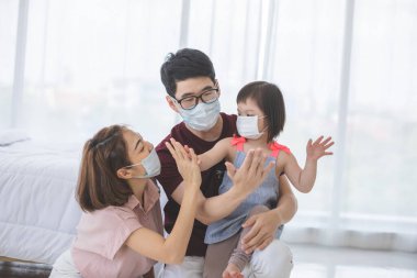Yüzünde tıbbi maskeler olan Asyalı aile, PM2.5 toz, duman, hava kirliliği ve COVID-19 'u önlemek için oturma odasında dururken kameraya bakıyor. Sağlık hizmetleri kavramı.