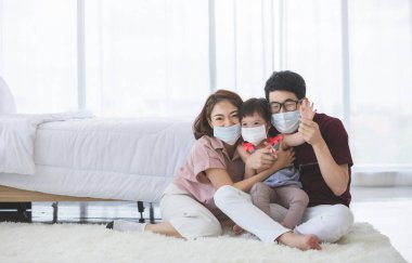 Yüzünde tıbbi maskeler olan Asyalı aile, PM2.5 toz, duman, hava kirliliği ve COVID-19 'u önlemek için oturma odasında dururken kameraya bakıyor. Sağlık hizmetleri kavramı.