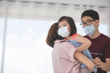 Yüzünde tıbbi maskeler olan Asyalı aile, PM2.5 toz, duman, hava kirliliği ve COVID-19 'u önlemek için oturma odasında dururken kameraya bakıyor. Sağlık hizmetleri kavramı.