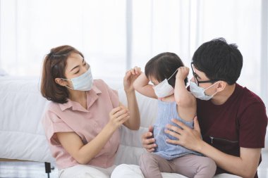 Yüzünde tıbbi maskeler olan Asyalı aile, PM2.5 toz, duman, hava kirliliği ve COVID-19 'u önlemek için oturma odasında dururken kameraya bakıyor. Sağlık hizmetleri kavramı.