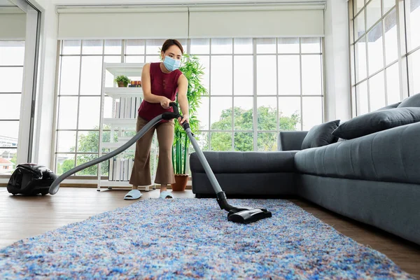 Beautiful young Asia woman wearing face mask for protect dust and using a vacuum cleaner while cleaning floor in the livingroom at home.Housekeeping occupation concept.