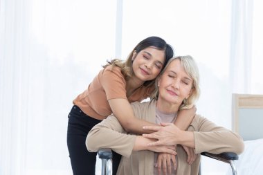 Anne ve evlatlık kızı birbirlerine tekerlekli sandalyede sarılıyorlar. Yatak odasının beyaz renginde samimiyet ve nezaketle. Alışılmadık görünüş, çeşitlilik, kalıtım konsepti..