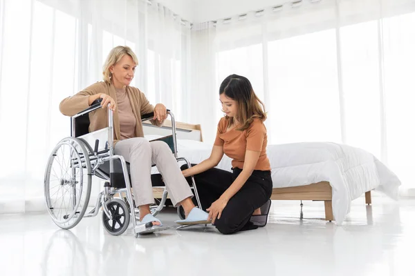 Mother and her adopted daughter take care her on a wheelchair with warmth and kindness in the bed room color white.Unusual appearance, diversity, heredity concept.