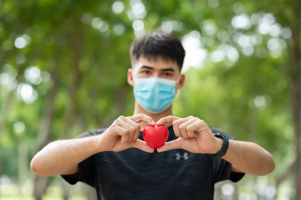 Young asian man in black shirt and protective masks against virus and air pollution,holding red heart make gesture for health care concept,environment green tree background.