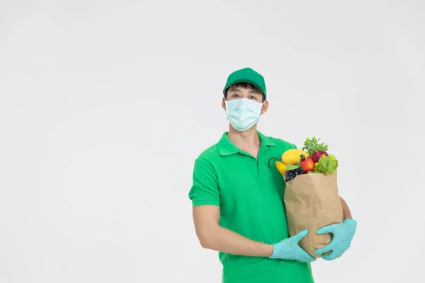顧客のために受信する新鮮な食品を渡す緑色の制服を着た顔のマスクを身に着けているスマートフードデリバリーサービスマンは オンライン注文で買い物をしています ロイヤリティフリーのストック写真