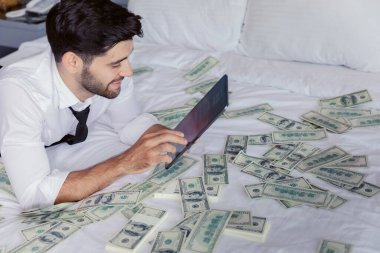 Tabletli mutlu genç işadamı etrafında dolarla yatakta dinleniyor. Başarı ve servet kavramı.
