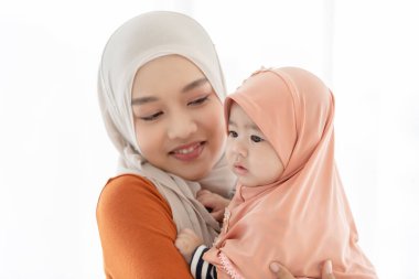 Müslüman bir anne ve küçük çocuk yatak odalarında. Anne başörtüsü takıyor ve bebeğini kucağında tutarken yatakta oturuyor ve ikisi de gülümsüyor..