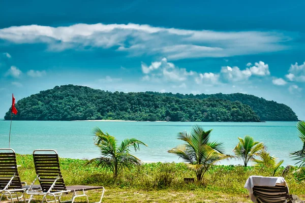 Small palm trees and deck chairs looking at a tropical island in the azure sea under the blue sky on the shores of the sandy beautiful exotic and stunning Cenang beach in Langkawi island, in Malaysia