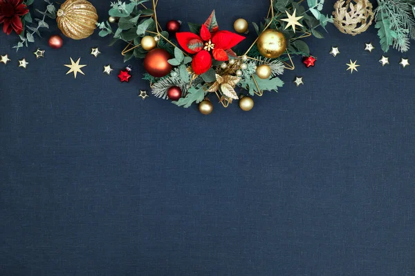 Decorative Christmas border, floral garland with eucalyptus, baubles, trinkets and red poinsettia. Red, green and golden Xmas decorations on classic blue linen background, copy-space.