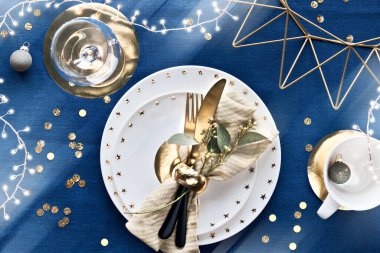 Beyaz tabak, altın çatal bıçak, yaldızlı geometrik metal ve kağıt dekorasyonlu bir Noel masası. Düz bir zemin, koyu mavi keten kumaş masa örtüsünün üstü. Xmas ışıkları çelenk.