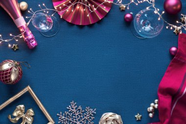 Altın ve pembe süslemeli Noel dekoratif kenarı. Şampanya şişesi, içki bardağı, kağıt deco, ıvır zıvır, şenlikli çelenk. Klasik mavi keten arkaplan üzerinde düz uzanma, kopyalama alanı..