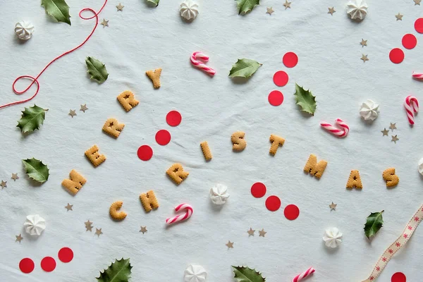 Doğal kutsal yapraklı çevre dostu Xmas dekoru. Beyaz tekstil yatıyordu. Kurabiyeden yapılmış mutlu Noeller mesajı. Xmas dekoru siluetleri kahverengi ve kırmızı kağıtlardan ve tatlı şekerlerden kesilmiş..