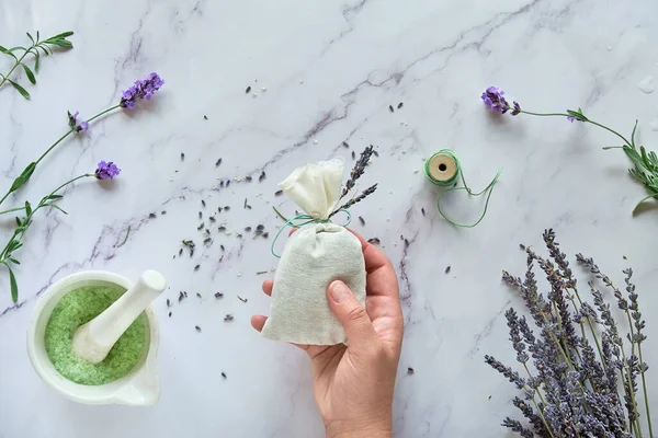 DIY handmade lavender sachets and sugar scrub. Hand holds linen bag with dry lavender flowers. Flat lay, top view on white marble background.