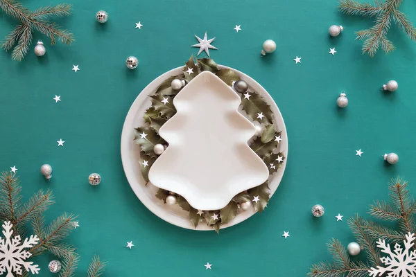 Christmas background in green and white. Xmas tree shape plate with copy-space on green mint textile tablecloth. Fir tree twigs, snowflakes, paper confetti. Holly leaves on round plate.