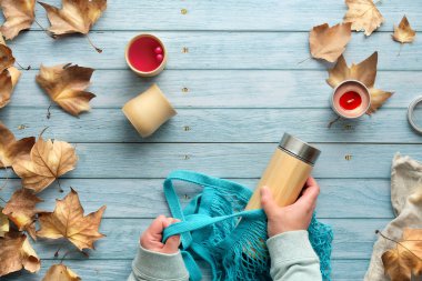 Ellerinde ağ torbası ve yalıtılmış bambu termos şişesi var. Sonbaharda sıfır ziyan çayı. Autumn 'un A metni. Bambu bardakları ve dağınık sonbahar yapraklarıyla solmuş ahşap masada yatıyordu..