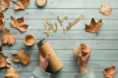 Sonbaharda sıfır atık içecek. Elleri çevre dostu yalıtılmış metal matara ve bambu kabı tutuyor. Sönük ahşap arka planda, dağınık sonbahar çınar yapraklarıyla yatıyordu..