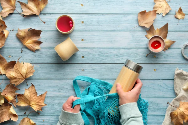 Ellerinde ağ torbası ve yalıtılmış bambu termos şişesi var. Sonbaharda sıfır ziyan çayı. Autumn 'un A metni. Bambu bardakları ve dağınık sonbahar yapraklarıyla solmuş ahşap masada yatıyordu..