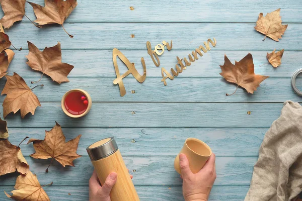 Elleri yalıtılmış metal matara ve bambu kabı tutuyor. Sonbaharda sıfır ziyan çayı. Düz, soluk, hafif tahtalar ve dağınık sonbahar yaprakları üzerinde yatıyordu..