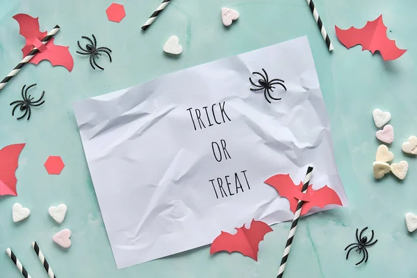 Trick or treat, text on white crumpled paper on green mint background. Flat lay, trendy Halloween background with checkered paper straws, flying bat silhouettes, sweet sugar hearts and spiders.