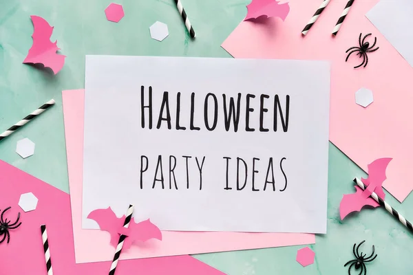 Text Halloween party ideas on layered paper background in mint green and pastel pink. Flat lay, Halloween party decor - hexagon confetti, drink straws, bats and spiders.