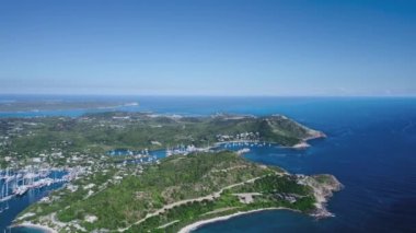 İnsansız hava aracı yoğun bitki örtüsüyle tepelik bir ada ve kuş bakışı Antigua ve Barbuda manzaralı beyaz yatlarla marina vuruyor. 