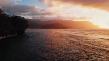 İnsansız hava aracı dalgaların üzerinde mor gün batımını, ufuktaki kara dağları Kauai, Hawaii, ABD 'de vuruyor.